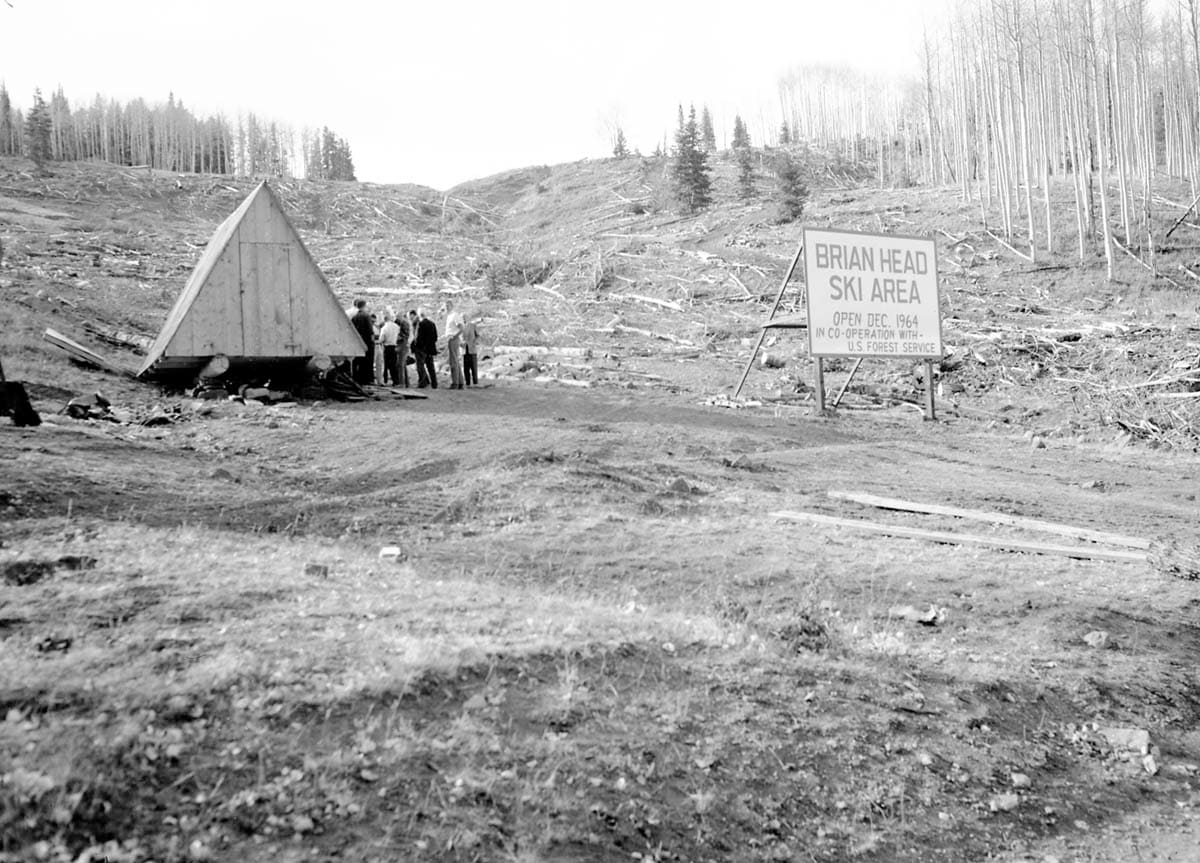 Black and white photograph of men standing at the Bear Flat ski development near Brian Head Peak in 1964, beside a sign announcing the ski area opening in December 1964