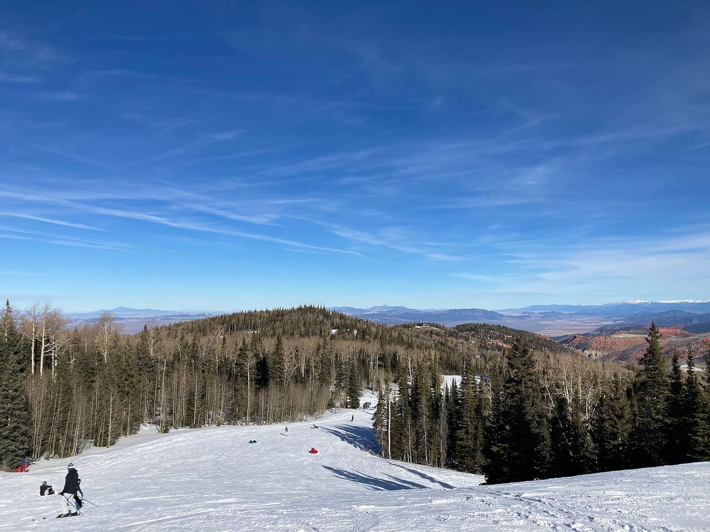 A groomed run at Brian Head with skiers, looking out over forested ridges toward red rock country and distant snow-capped peaks