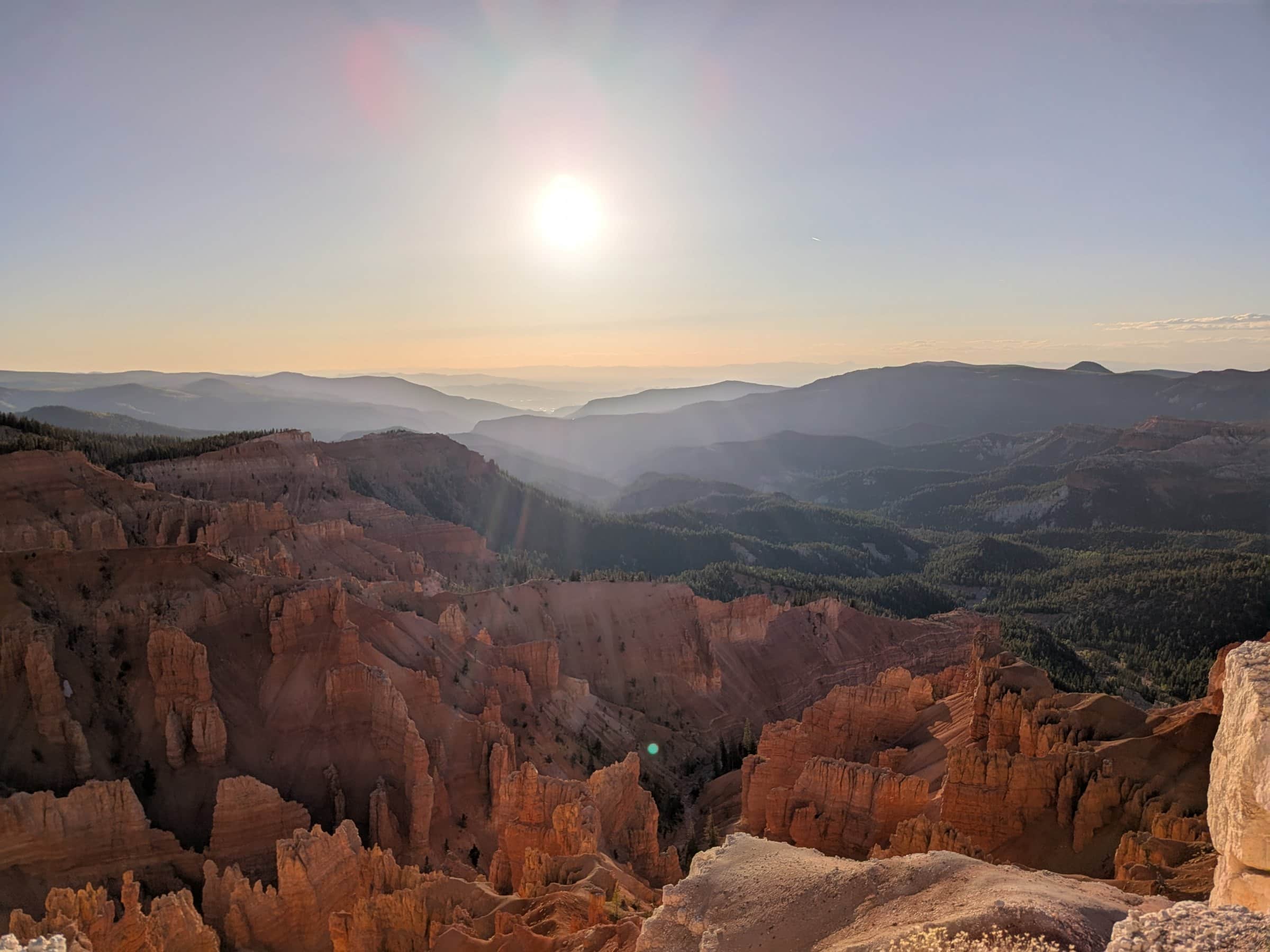 The Cedar Breaks amphitheater in evening light, red and orange hoodoos in the foreground and forested ridges fading toward the sun