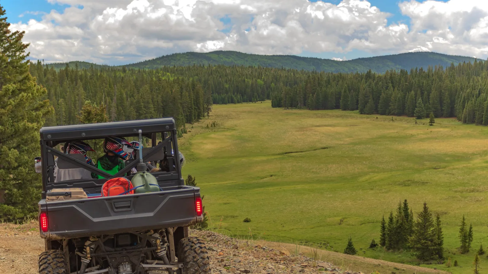 Un UTV side-by-side en un camino de grava sobre una pradera verde de montaña bordeada de pinos, con nubes altas formándose sobre las colinas boscosas de la meseta del Markagunt
