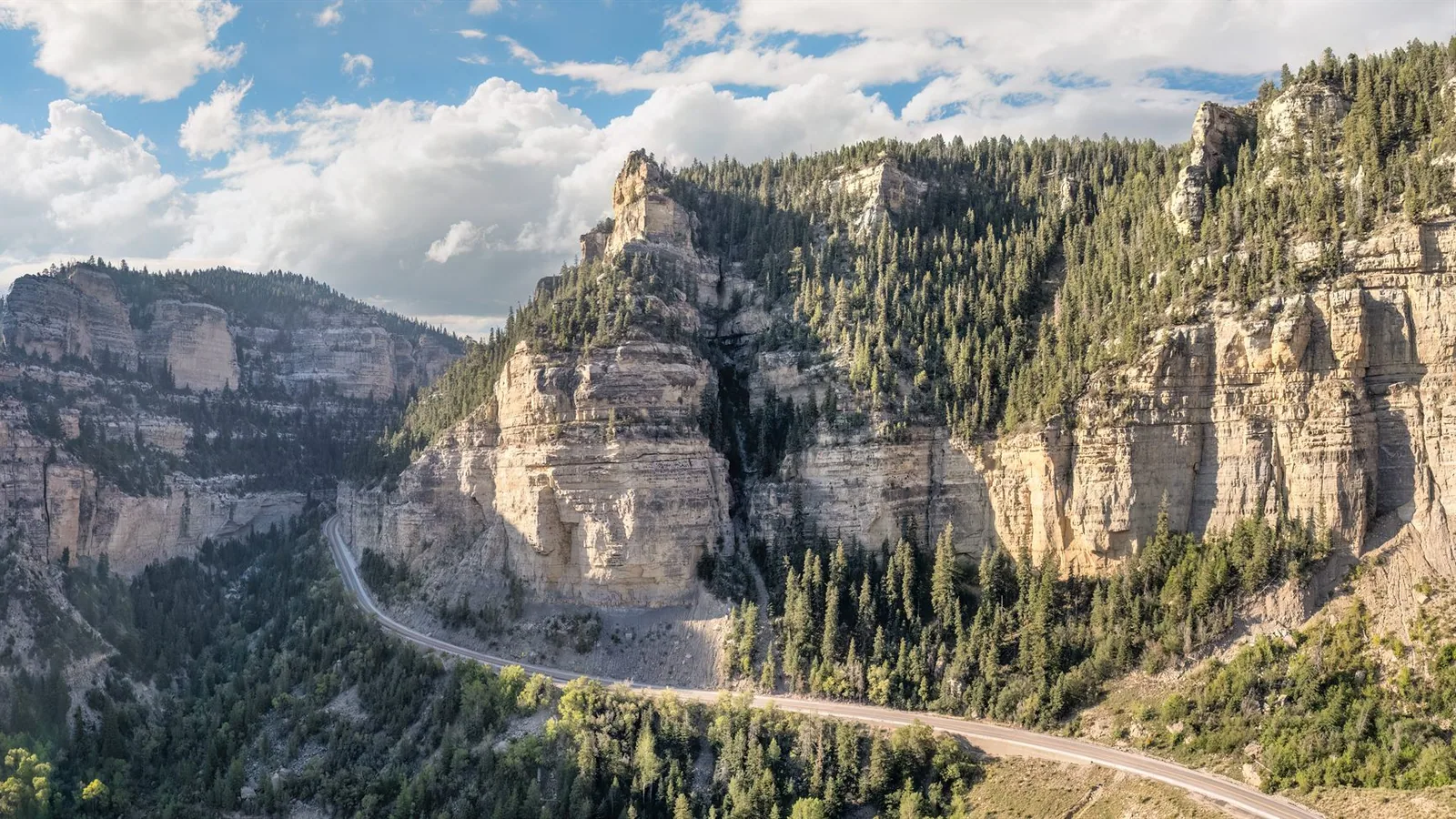 La Highway 14 serpente dans Cedar Canyon sous des falaises stratifiées roses et crème coiffées de conifères sombres, hauts nuages d'été.