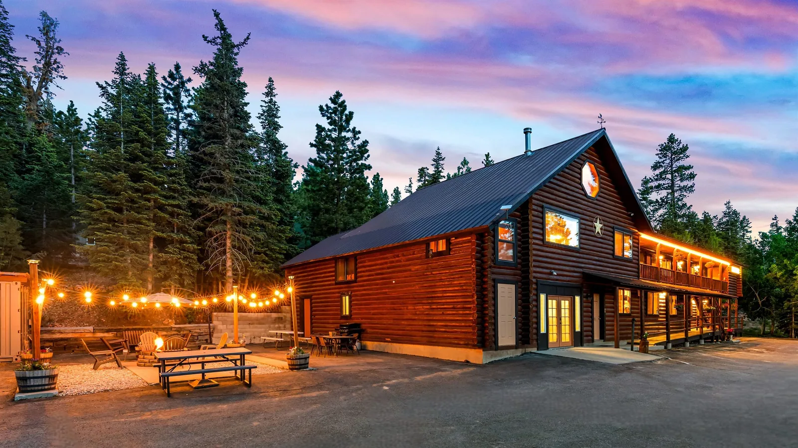 Bâtiment en rondins du Duck Creek Village Inn au crépuscule, guirlandes lumineuses au-dessus du foyer extérieur et ciel rose et violet au-dessus des pins