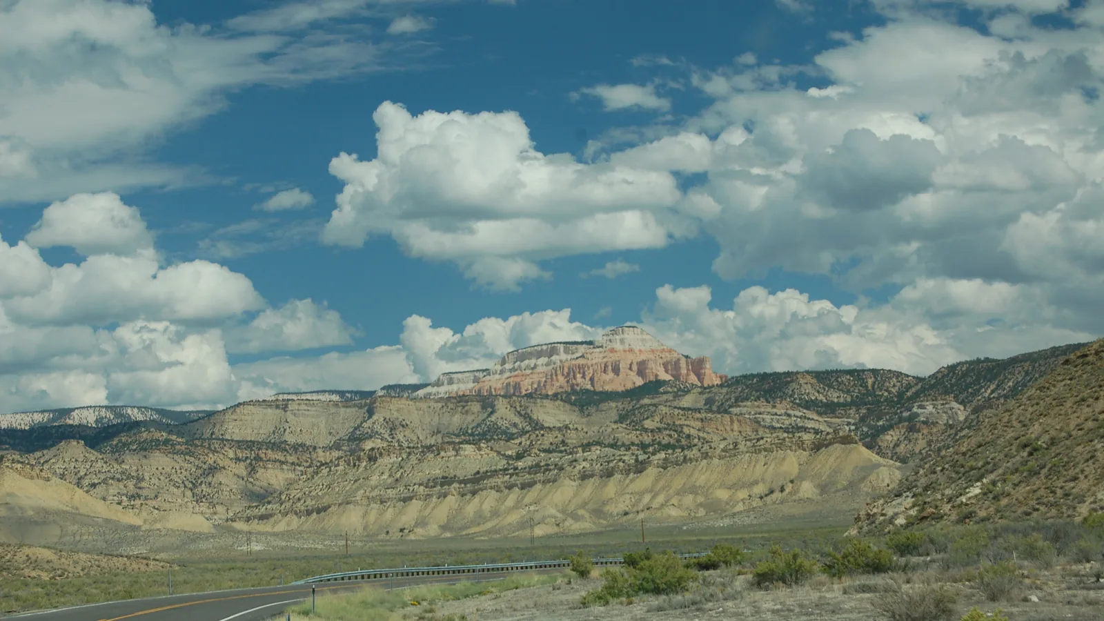 Scenic Byway 12 curves past pale badlands toward the pink and white cliffs of Powell Point under a sky of white clouds