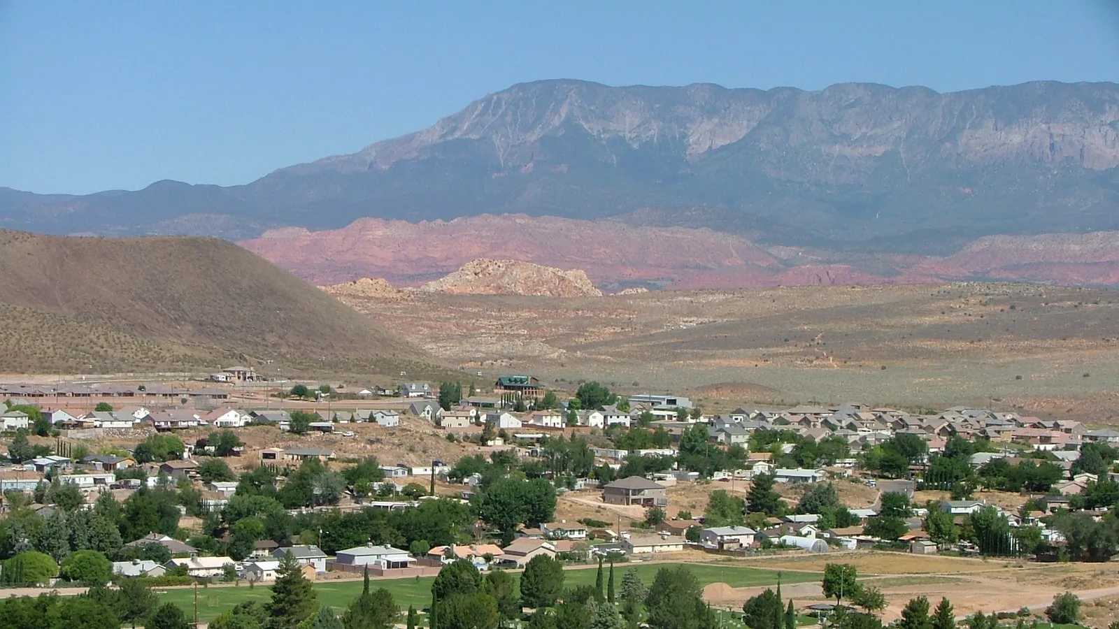 犹他州飓风镇的民居与绿树，背后是红岩山丘和灰色高山山脊，夏日晴空万里