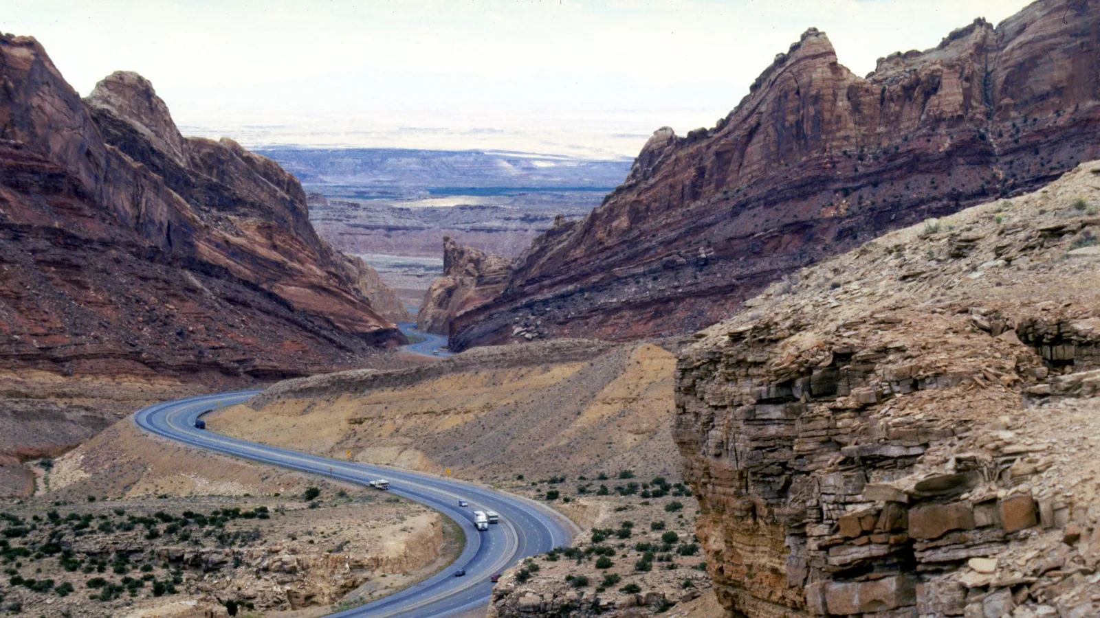 Interstate 70 curves through a red rock canyon in the San Rafael Swell with trucks and cars on the road and desert flats beyond
