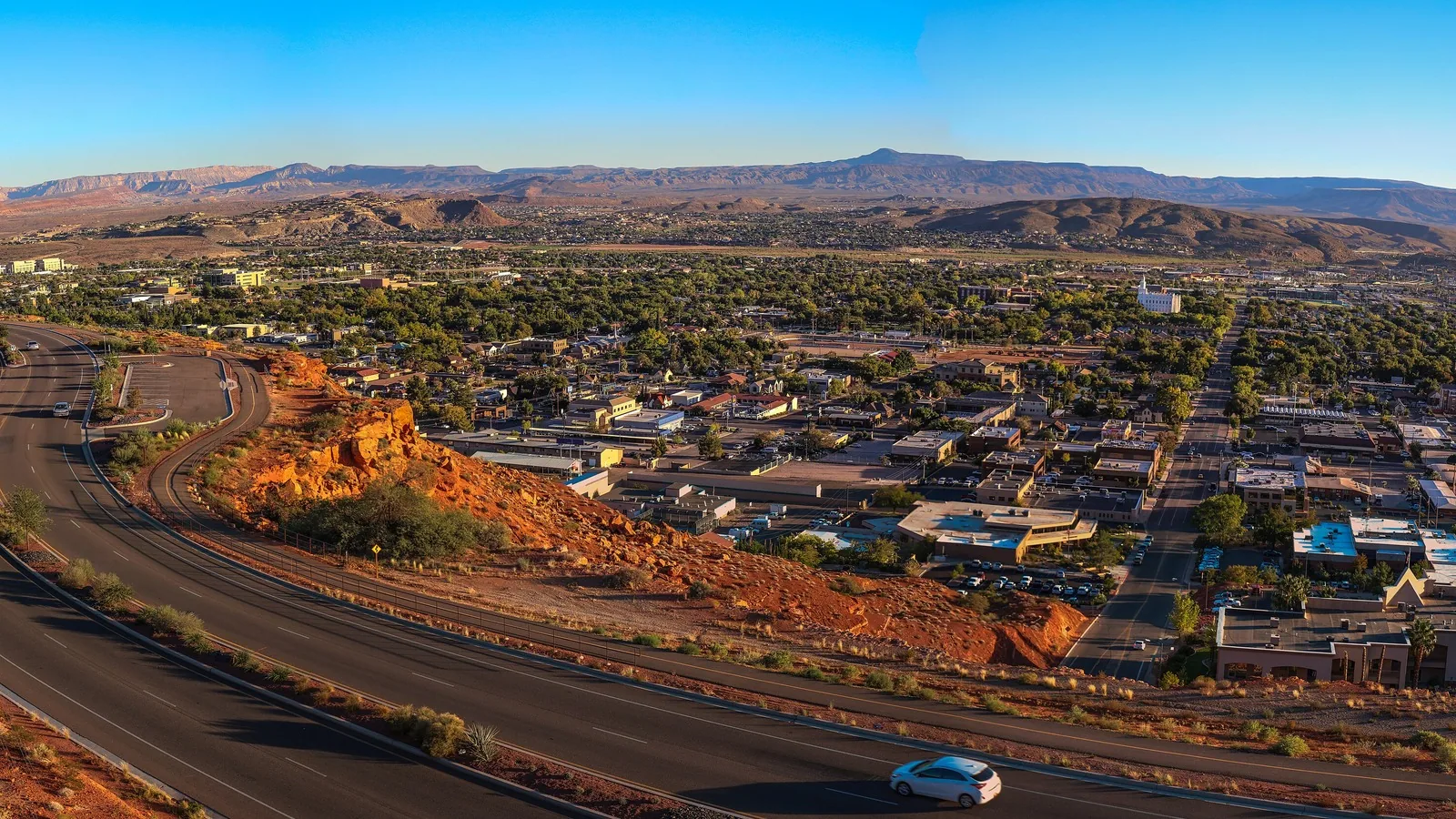 Downtown St. George, Utah at evening with a curving road and red rock hill in the foreground and the Pine Valley Mountains on the horizon