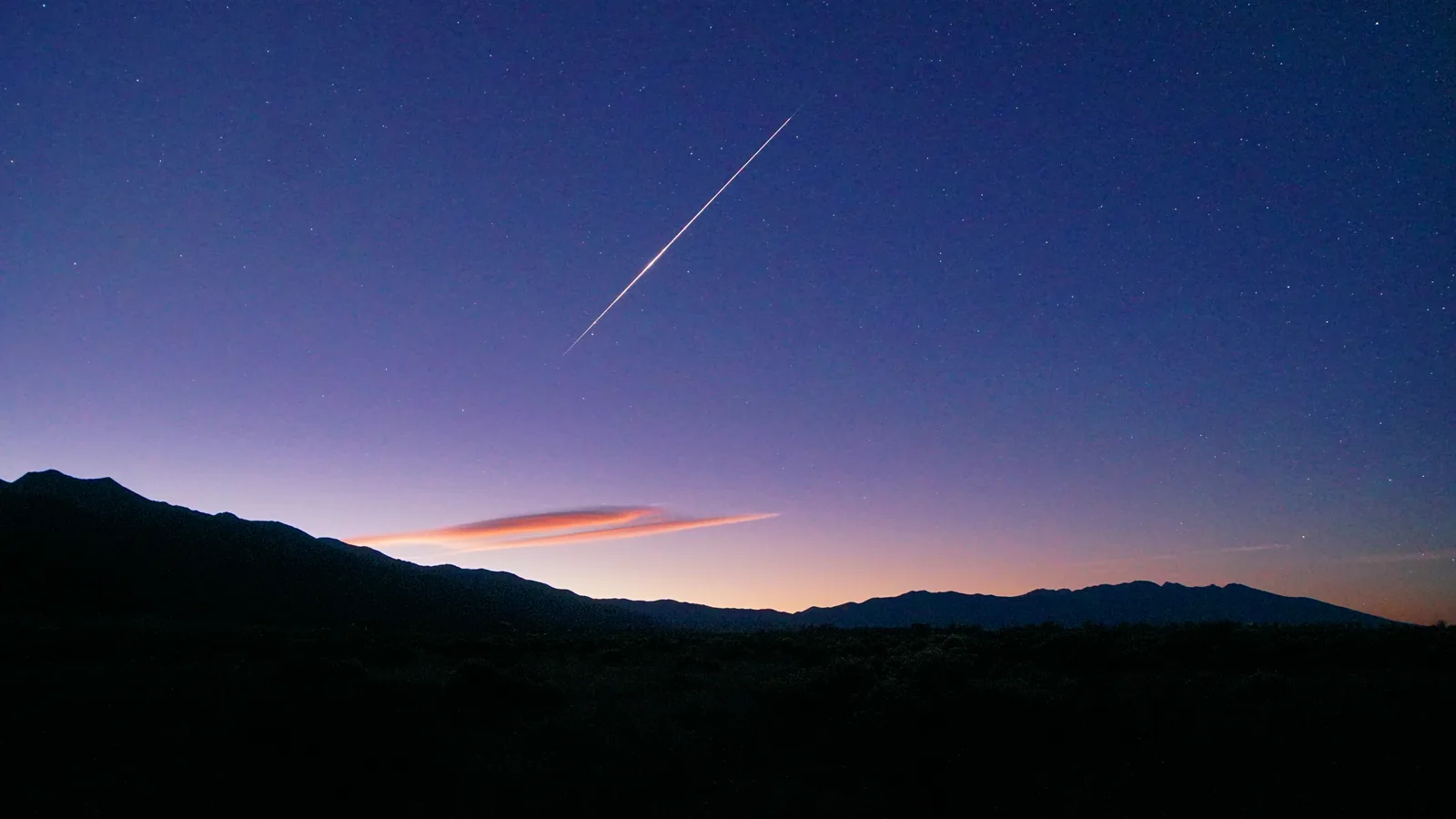 Un solo meteoro brillante cruza un cielo azul profundo antes del amanecer, sobre una cresta de montaña oscura y una fina nube lenticular rosada