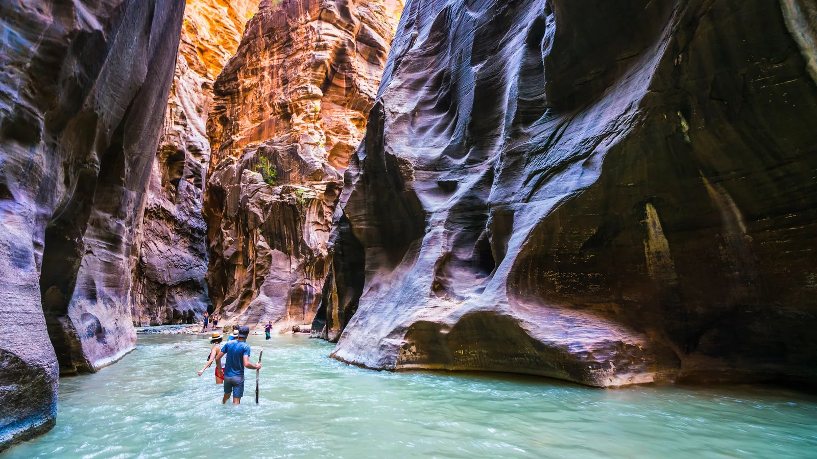 Deux randonneurs remontent un courant peu profond aux reflets verts entre de hautes parois de grès striées dans un canyon étroit, lumière chaude en haut de la roche.